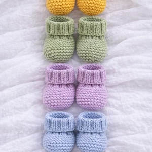 May include: A collection of five pairs of handmade baby booties in various colors. The booties are knitted in yellow, green, lavender, light blue, and brown. The booties are arranged in rows on a white, textured surface.