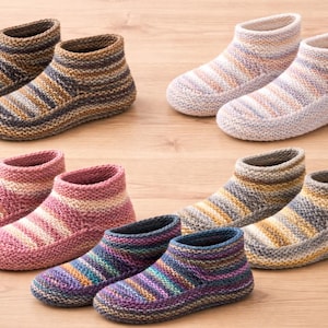 Op de afbeelding: Een verzameling gebreide pantoffelboots in verschillende kleurencombinaties. De laarzen hebben gestreepte patronen in tinten bruin, roze, paars en blauw. Het schoeisel is gerangschikt op een lichtbruin houten oppervlak.