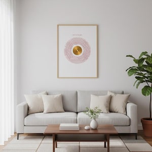 Può includere: Una stampa incorniciata con un design circolare rosa e oro, con l'etichetta "ARTIST NAME". L'opera d'arte è appesa sopra un divano grigio chiaro con cuscini beige. Un tavolino in legno e un tappeto fantasia completano il soggiorno.
