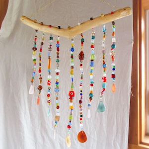 Può includere: Una giostrina sospesa con un ramo di legno chiaro a forma di V appeso a una sottile catenella dorata. Molteplici fili di perline colorate in vetro, plastica e pietra di varie forme e dimensioni pendono dal ramo, terminando con ciondoli piatti.