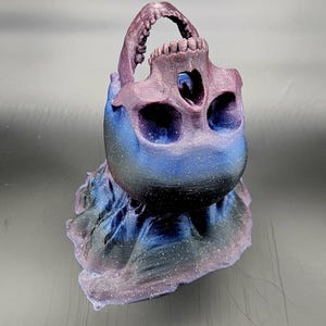 Puede incluir: Una escultura de cráneo impresa en 3D con un acabado tipo galaxia en tonos de morado, azul y negro. El cráneo está inclinado hacia atrás, revelando el interior del cráneo.