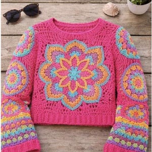 Könnte beinhalten: Ein leuchtend rosa Häkelpullover mit einem großen Blumendesign in der Mitte und auf den Ärmeln. Das Blumendesign weist Farbtöne von Blau, Orange und Gelb auf. Der Pullover hat lange Ärmel und einen runden Ausschnitt. Eine Sonnenbrille ist oben links zu sehen.