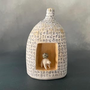 May include: A beige ceramic sculpture shaped like a small house with a textured roof. The exterior is covered in black printed letters. A rectangular opening reveals a small white animal figurine with a spotted ball balanced on its back.