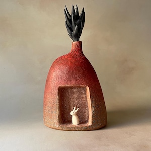 May include: A dome-shaped ceramic sculpture with a textured red and orange finish. The piece features a rectangular recessed opening containing a small, smooth white rabbit figurine. A black, spiky, plant-like form extends from the top.