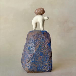 May include: A ceramic sculpture featuring a white bear standing on a large, textured brown rock with blue swirling patterns. A small, round, speckled brown stone is balanced on the bear's back. The piece is crafted from clay.