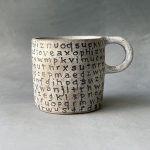 May include: A speckled ceramic mug with a rounded handle. The mug's exterior is covered in a random pattern of black letters. The mug has a neutral color palette, with a light beige base and black lettering.