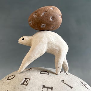 Op de afbeelding: Een grillige sculptuur van een wit wezen dat een bruine steen op zijn rug balanceert. Het wezen staat op een ronde basis met geëtste letters. De steen heeft kleine, gebeeldhouwde inkepingen. De achtergrond is grijs.