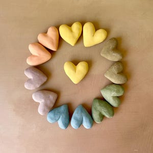 May include: A circular arrangement of heart-shaped objects in pastel colours. The hearts are in shades of yellow, peach, blue, and green, set against a neutral background. The arrangement suggests a decorative or artistic theme, possibly for Valentine's Day.