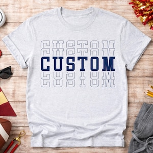 May include: A light gray t-shirt with the word "CUSTOM" in navy blue, stacked vertically. The word is also outlined in a lighter blue, creating a layered effect. The shirt is on a wooden surface.