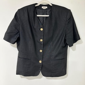 May include: A black short-sleeved blazer featuring a V-neckline and four light-coloured, speckled buttons down the front. The garment includes two horizontal welt pockets at the waist and is displayed on a white hanger against a plain background.