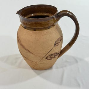 May include: A stoneware pitcher with a tan, unglazed body and a dark brown glazed rim and handle. The exterior features etched, dark brown leaf and stem designs. The pitcher has a wide, rounded base, a curved handle, and a pouring spout.