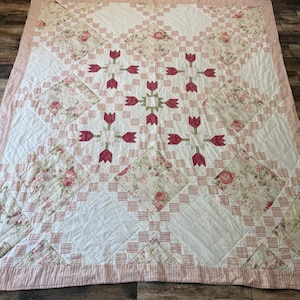 May include: A patchwork quilt featuring a repeating pattern of white squares, floral print squares with pink roses, and tulip motifs in dark pink and green. The quilt is bordered by a thin pink and white striped fabric trim along the edges.