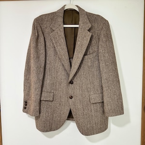 May include: A brown blazer featuring a classic herringbone pattern, notched lapels, and two front buttons. The garment includes two flap pockets at the waist, a single chest pocket, and three small buttons on each sleeve cuff, with a solid brown lining.