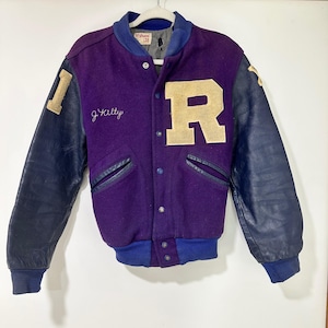 Pode incluir: Uma jaqueta universitária roxa com mangas de couro azul-escuro e punhos canelados. A frente apresenta uma letra R grande na cor creme, o nome bordado J. Kilty e uma etiqueta da marca Wilson com o tamanho 38. Fecho com botões de pressão.