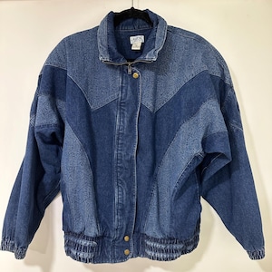 May include: A blue denim jacket featuring a chevron-patterned panel with an embossed paisley design. The jacket has a high collar, a front zipper closure, two gold-tone metal buttons at the hem, and an elasticized waistband and sleeve cuffs.