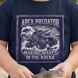 May include: Navy blue t-shirt featuring a shark-themed monster truck graphic with the text "APEX PREDATOR" and "MAKING WAVES IN THE ARENA". The shirt is worn by a young child holding a toy shark.