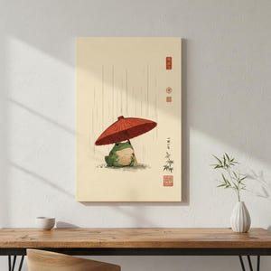 以下が含まれることがあります： クリーム色の背景に、赤い傘をさして雨の中に座る緑色のカエルの絵が描かれた長方形のウォールアートプリント。雨を表す縦線、竹のイラスト、赤い日本語の文字と印章がデザインに含まれています。