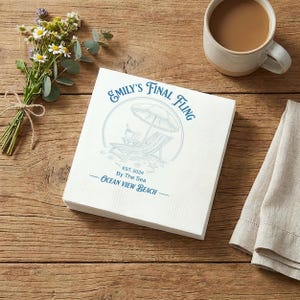 May include: A stack of white paper napkins with blue text and a beach-themed illustration. The text reads "Emily's Final Fling" and "Ocean View Beach". A cup of coffee, flowers, and a linen napkin are also visible.