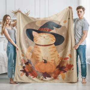 Könnte beinhalten: Eine beige Fleecedecke mit einer grafischen Illustration einer orangefarbenen getigerten Katze, die einen schwarzen Hexenhut trägt. Die Katze sitzt hinter einem großen orangefarbenen Kürbis, umgeben von herbstlichen Blättern in Rot, Braun und Gold.