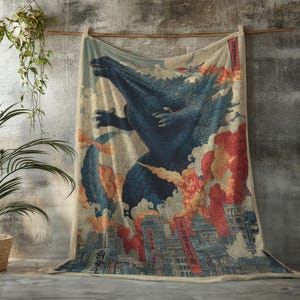 May include: A beige blanket featuring a blue Godzilla illustration in a vintage Japanese art style. The design includes a city scene with red and orange flames, and Japanese text. The blanket is hanging on a wooden rod.