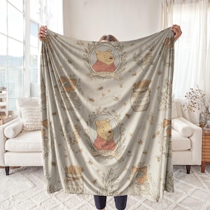 May include: A beige blanket featuring a repeating pattern of Winnie the Pooh in a circular frame, jars of honey, bees, and floral accents. The blanket has a soft, plush texture and is held up by a person.