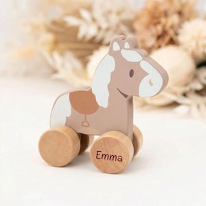 Op de afbeelding: Een houten trekpaard in tinten beige, bruin en wit. Het paard heeft een zadel en een witte manen en staart. De naam "Emma" staat op een van de houten wielen. Het speelgoed is ontworpen voor jonge kinderen.