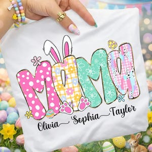 May include: White t-shirt with the word "MAMA" in colorful, patterned letters, including pink polka dots, yellow flowers, and blue and pink plaid. The names "Olivia", "Sophia", and "Taylor" are written below.