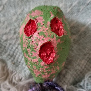 May include: A green and pink doll head with red painted eyes and mouth. The doll head is attached to a body made of yarn.