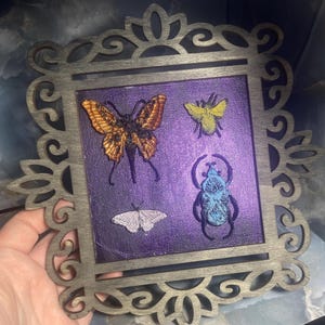May include: A decorative wooden frame with a purple background featuring detailed insect art. The insects include a butterfly, a bee, a moth, and a beetle. The frame has an ornate, cut-out design.