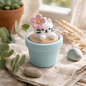 Pode incluir: Um vaso de cerâmica azul claro com uma figura decorativa de gato. O gato branco tem detalhes faciais pretos e uma flor rosa na cabeça. A figura está sobre uma base bege dentro do vaso, com pequenas pedras e uma planta suculenta no fundo.