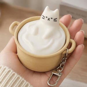 Pode incluir: Um chaveiro extravagante com uma figura de gato branco aparentemente submersa em uma substância cremosa dentro de uma pequena panela bege. O gato tem detalhes faciais pretos. Uma corrente e um fecho prateados estão anexados.