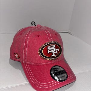 Peut inclure: Casquette de baseball rouge avec le logo des 49ers de San Francisco. La casquette présente une visière incurvée, des coutures blanches et un logo décoratif avec des strass. Le chapeau réglable porte une étiquette "9TWENTY".