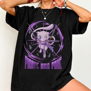 May include: A black short-sleeve t-shirt featuring a graphic of a light purple creature with a long tail, holding two swords. The design includes a circular purple energy effect and the text PIRATE KING POKE in purple block letters at the bottom.