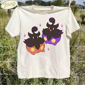 May include: An ivory short-sleeve t-shirt featuring a graphic of two dark-colored, pumpkin-like creatures with yellow eyes. One creature has an orange base and the other has a purple base. Small purple star-like accents surround the figures.