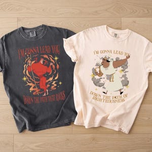 May include: Two t-shirts with graphic designs. One is dark gray with a red devil illustration and the text "I'M GONNA LEAD YOU DOWN THE PATH THAT ROCKS." The other is light pink with a cartoon illustration and the text "I'M GONNA LEAD YOU DOWN THE PATH OF RIGHTEOUSNESS."