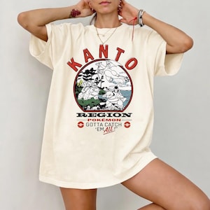 Puede incluir: Camiseta color crema con un diseño circular. El diseño presenta el texto "KANTO REGION POKÉMON GOTTA CATCH 'EM ALL!" e ilustraciones de personajes Pokémon en un paisaje, con detalles en rojo y negro.