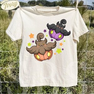 May include: An ivory short-sleeve t-shirt featuring a graphic of two pumpkin-like creatures with dark brown and black hair, orange and purple bases, and glowing yellow eyes, surrounded by small colorful stars in orange, purple, and lime green.