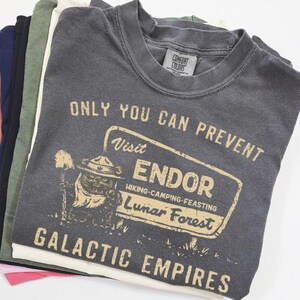 May include: Dark grey t-shirt featuring the text "ONLY YOU CAN PREVENT Visit ENDOR Lunar Forest GALACTIC EMPIRES." The shirt has a vintage-style graphic of a creature with a hat and shovel. The shirt is folded with other coloured shirts.