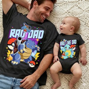 Puede incluir: Camiseta negra con un gráfico de "Rad Dad" con una tortuga de dibujos animados azul y una bola roja y blanca. Un bebé con un body negro con un gráfico a juego que dice "Rad Like Dad" también está en la imagen.