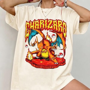 May include: An off-white short-sleeve t-shirt featuring a large graphic print of the orange dragon-like creature Charizard. The design includes the text CHARIZARD in yellow and red, flames, and three small red and white spherical Poké Balls.
