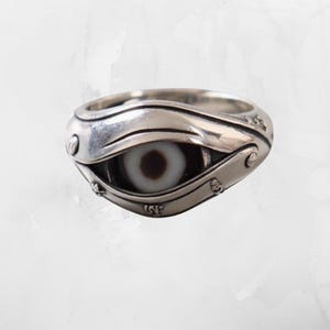 May include: A silver ring featuring a central eye motif with a white and dark brown stone inlay. The metal band is sculpted with layered, eyelid-like curves and small, embossed decorative details along the sides, creating a dimensional appearance.