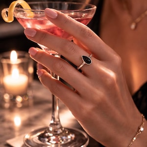 Puede incluir: Un anillo de plata con una piedra negra en forma de lágrima se lleva en un dedo. Se sostiene un cóctel con una rodaja de limón en un vaso. También se ven una pulsera y un collar de oro.