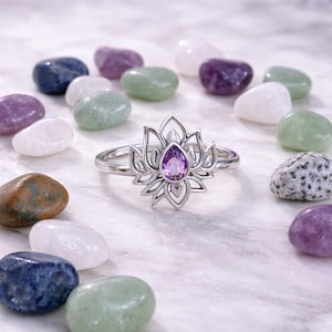 May include: A silver ring featuring a lotus flower design with a teardrop-shaped purple gemstone at its center. The ring is surrounded by colorful polished stones in shades of purple, green, blue, and white.