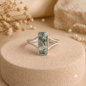 May include: A silver ring featuring a rectangular moss agate gemstone with green dendritic inclusions. The band has a split design that connects to the sides of the stone setting, creating a clean and balanced look for this piece of jewelry.