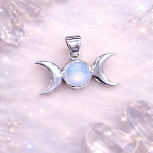 Pode incluir: Um pingente prateado com um design de lua tripla. Apresenta uma pedra central circular, ladeada por luas crescentes. O design é definido contra um fundo macio e brilhante, sugerindo um tema celestial.