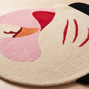 May include: A round rug with a whimsical design. The rug features a light pink, off-white, red, orange, and black color scheme. The rug's design resembles a cartoon character's face, with red lines and black ear-like shapes.