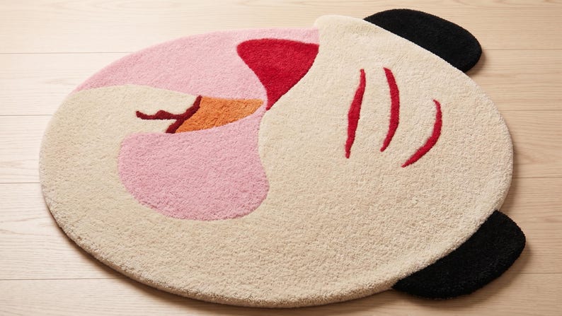 May include: A round rug with a whimsical design. The rug features a light pink, off-white, red, orange, and black color scheme. The rug's design resembles a cartoon character's face, with red lines and black ear-like shapes.
