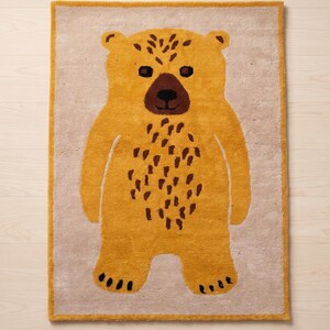 May include: A rectangular rug featuring a stylized bear design. The bear is primarily mustard yellow with brown accents on the face and body. The rug has a light beige border and background, suitable for a child's room.