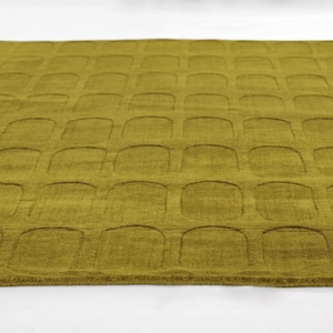 Puede incluir: Primer plano de una alfombra verde oliva texturizada. La alfombra presenta un patrón repetitivo de rectángulos redondeados, creando un diseño similar a una cuadrícula. La superficie de la alfombra parece suave y afelpada, con un brillo sutil.