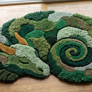 May include: A tufted rug shaped like a sleeping dragon, featuring a textured surface with various shades of green, including forest, olive, and mint. The dragon has two prominent golden-brown horns and a large, coiled tail in the center.